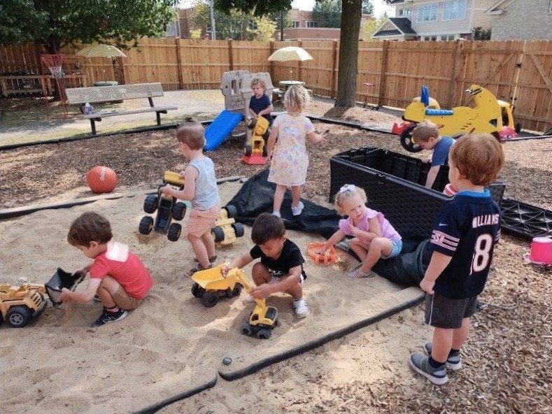 A group of children playing with trucks in the sandbox on the playground.