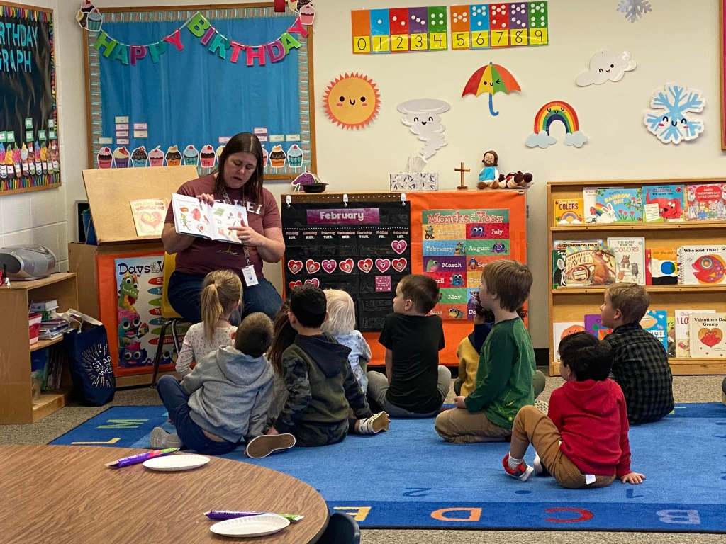 Classroom photo of children, facing away from the camera, and listening to a teacher read at story-time.