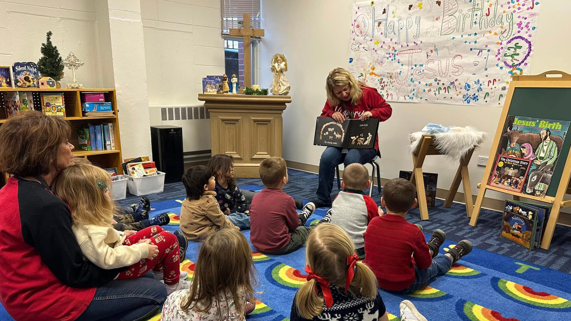 Children, facing away from the camera, listening to a book about the birth of Jesus in the Bible Study room.